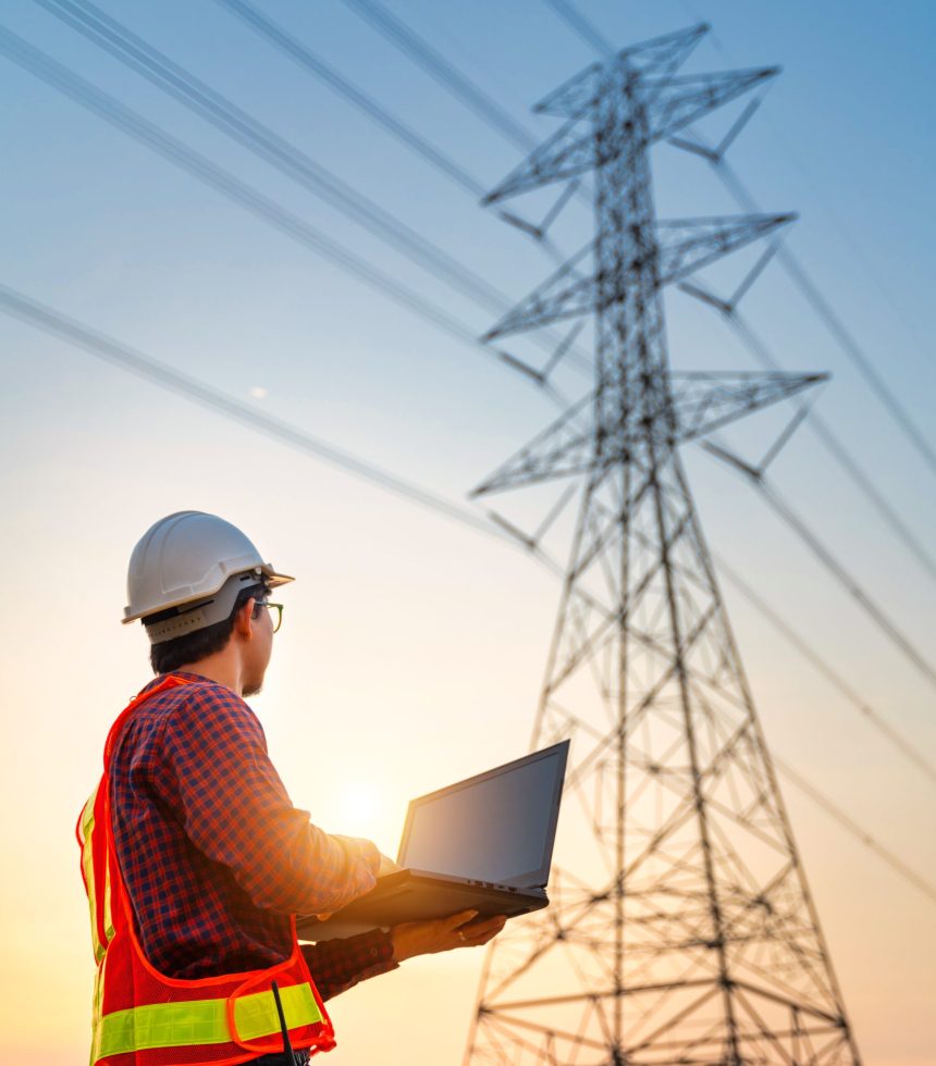 Asian electrical engineers checking location using a notebook computer standing at a power station to view the planning work by producing electrical energy at high voltage electrodes. Vertical image.